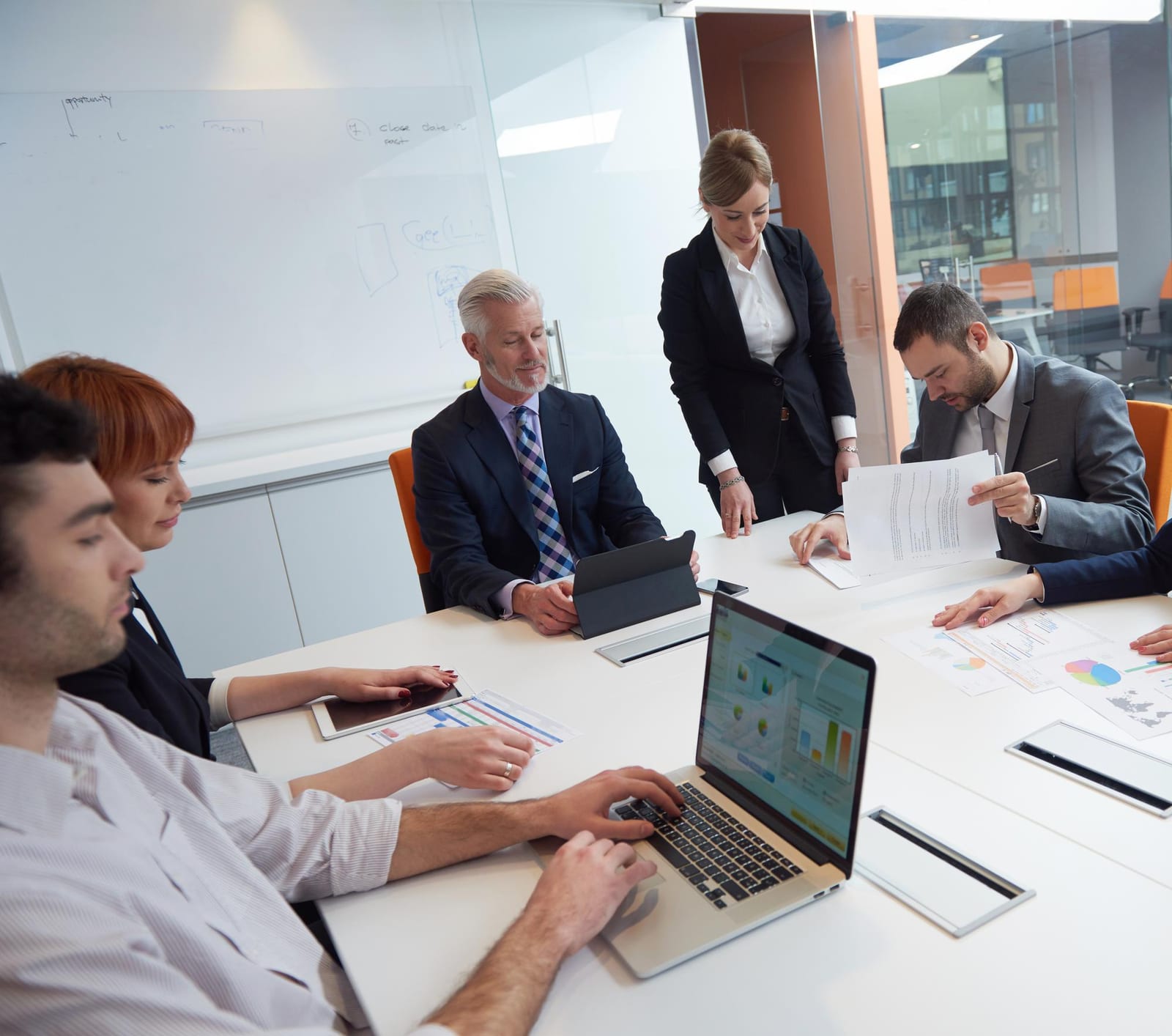 Sechs Kolleginnen und Kollegen in Geschäftskleidung besprechen sich an einem weissen Tisch mit Laptop, Tablet und Unterlagen vor einem Whiteboard.